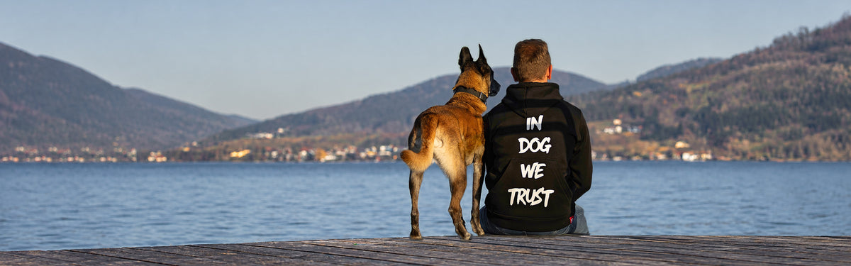 Frano sitzt mit einem Malinois am See und trägt den schwarzen IQ Hoodie „In Dog We Trust“.